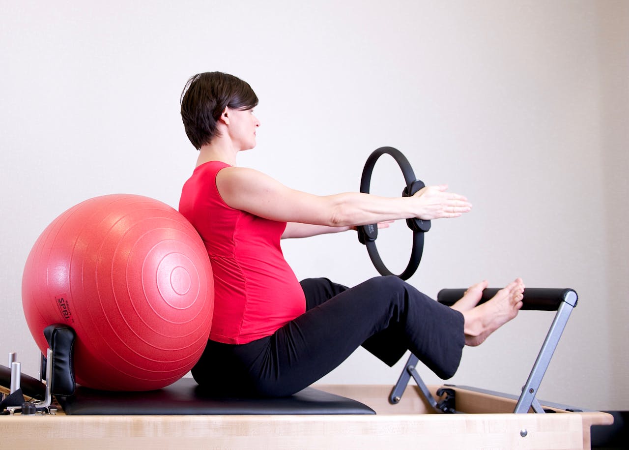 our-stories A pregnant woman in a red top practicing Pilates using a fitness ring and exercise ball in a gym setting.